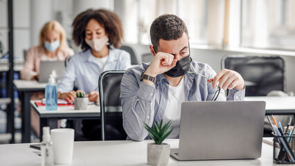 Tired and overworked. Focus on man in protective mask rubs his eyes with hand, behind sit female colleagues at workplaces with laptops and antiseptics in office interior during outbreak of covid-19