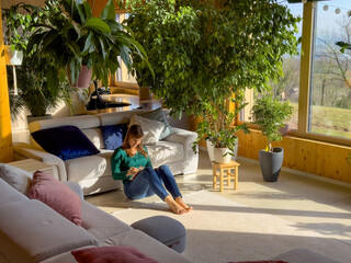 Young woman leans on a white sofa while sitting on the floor, completely immersed in a book. She...