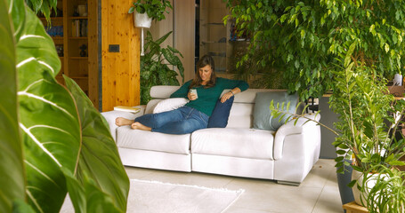 Young lady lounges comfortably on white couch, focused on her smartphone. Filled with lush indoor...