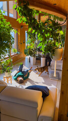 Dog owner lies on living room floor of a sunlit living room, smiling as she plays with her cute dog. Relaxed and playful moment between woman and pet, surrounded by lush plants and warm wood accents.