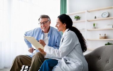 Obraz premium Young doctor asking senior impaired male patient in wheelchair to sign insurance policy at home. Handicapped elderly man putting his signature under surgery consent form, reading medical document