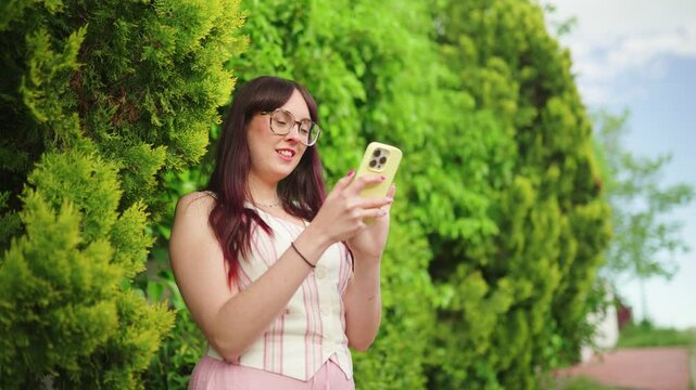 Mujer de pie sonriendo mientras escribe en su smartphone en un entorno eco natural frente a plantas y cielo azul