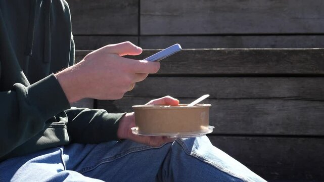 Close-up of a man eating healthy takeaway food and holding a smartphone in his hand, chatting with friends, scrolling through the news feed