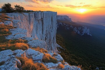 Sunset over towering white cliffs with green forest below, warm light illuminating rocky ledge and sparse trees under a partly cloudy sky