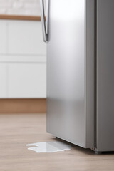 Silver refrigerator with a water puddle beneath it on a wooden floor. Appliance leak issue indoors
