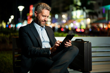 Plakat Businessman working on tablet outdoors. Businessman with tablet outdoor. Businessman with a tablet on night street. Businessman using tablet, sit on bench. Night business.
