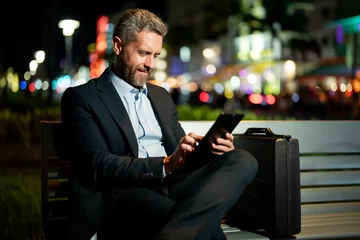 Gordijnen Businessman working on tablet outdoors. Businessman with tablet outdoor. Businessman with a tablet on night street. Businessman using tablet, sit on bench. Night business. © Volodymyr