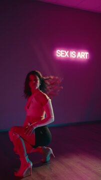 A brunette woman dances in a dimly lit studio with pink and blue neon lights in the background. She wears a white top, black shorts, and high heeled white boots. Young Woman Dancing Sensually