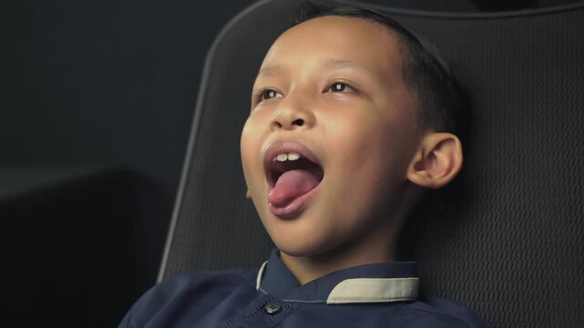 A little boy opening her mouth wide to show her throat. Child is sitting with wide open mouth in a black chair, with a tongue stuck out