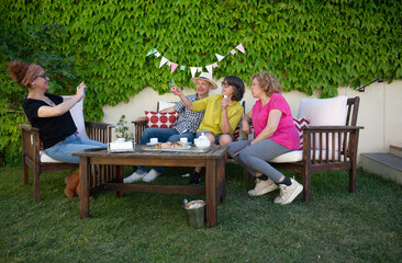 Middle-aged friends sitting outdoors in the backyard sharing coffee and pastries while taking...