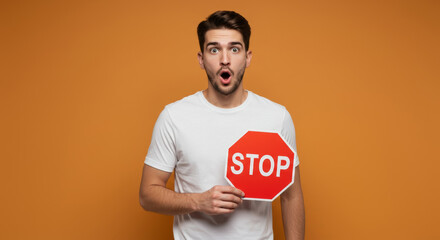 Young man in a white t-shirt holding a stop sign, surprised expression in front of a plain orange background. Adult male model holding a stop sign in studio setting. Casual wear.