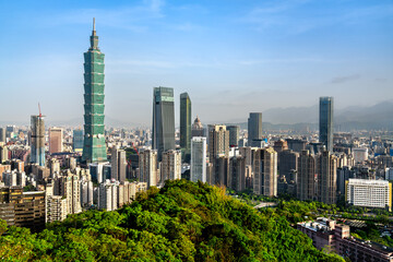 The skyline of Taipei, Taiwan, features a mix of modern skyscrapers and dense city buildings, rising behind a lush green hilltop and set against a bright blue sky with distant mountain ranges.
