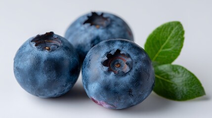 Obraz premium Three ripe blueberries with a single green leaf are showcased in a macro shot against a white background, emphasizing their vibrant colors and fresh appearance