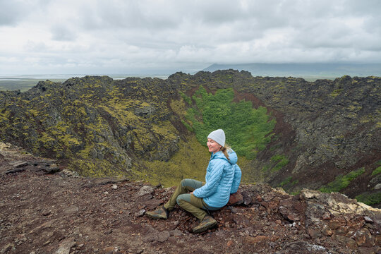 Woman in jackets sitting on Eldborg volcanic crater edge in Iceland