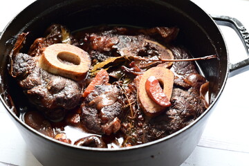 Close-up of a rustic gourmet dish featuring slow-braised beef shank with bone (osso buco style), served on black pot ,white background
