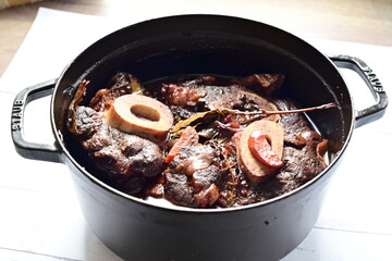 Close-up of a rustic gourmet dish featuring slow-braised beef shank with bone (osso buco style), served on black pot ,white background