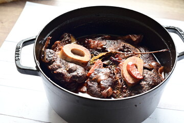 Close-up of a rustic gourmet dish featuring slow-braised beef shank with bone (osso buco style), served on black pot ,white background