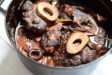 Close-up of a rustic gourmet dish featuring slow-braised beef shank with bone (osso buco style), served on black pot ,white background