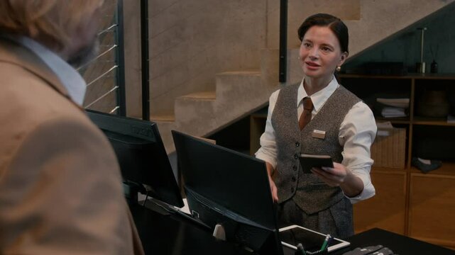 Over the shoulder view of female hotel receptionist talking to guest and reviewing his document behind counter during check in process