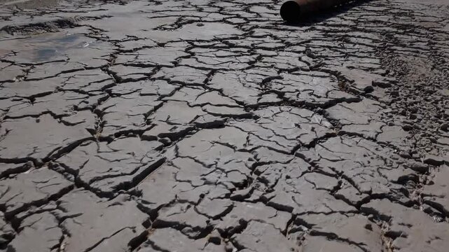 Parched riverbed revealing deep soil cracks, abandoned rusty industrial pipe symbolizing environmental degradation from prolonged drought and climate change