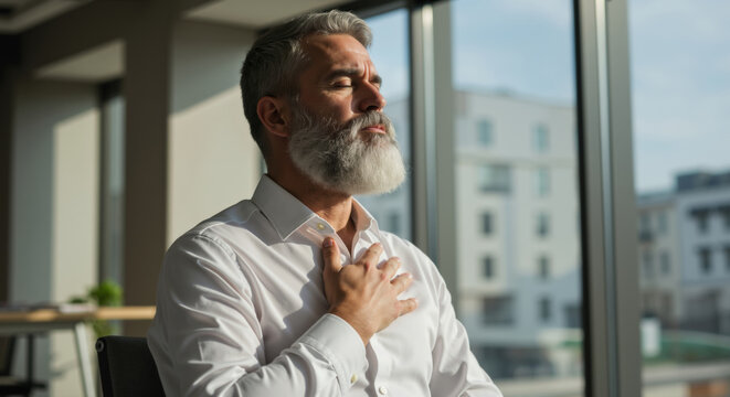 Senior man with white beard and closed eyes placing hand on heart in bright office. Mindful business practice for gratitude expression and emotional wellbeing in corporate wellness