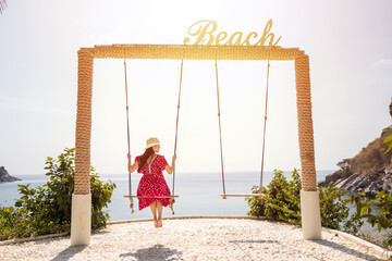 back view of Asian woman in red maxi dress sitting on a swing with a sign "Beach" on top, sea view at the background. happy summer travel concept