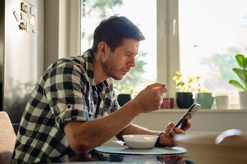 Man in plaid shirt eating oatmeal porridge from white bowl while using smartphone. Male looking at phone during breakfast. Concept of social media addiction, distraction, and digital dependency