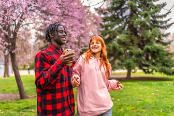 Fototapeta premium Young couple holding hands and using smartphone in blooming park