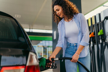Young woman refueling her vehicle at a self service gas station, holding the fuel pump nozzle and waiting for the tank to be full during a sunny summer day