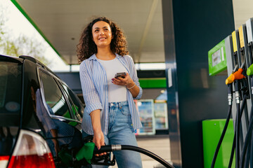 Happy young woman refueling her car at a gas station, holding a smartphone in one hand and a fuel pump in the other, enjoying the convenience of modern technology