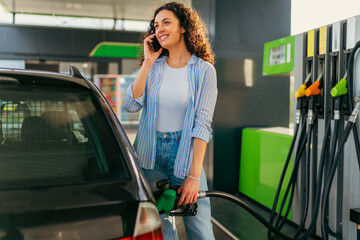 Smiling woman refueling her car at a gas station while chatting on her mobile phone, embracing the...