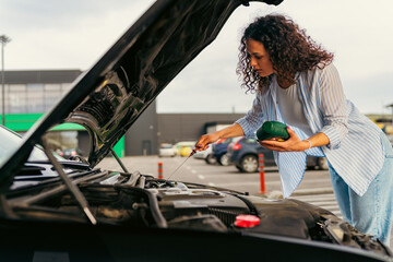 Young woman checking the oil level in her car engine using a dipstick, holding a bottle of motor oil, performing routine maintenance in a parking lot