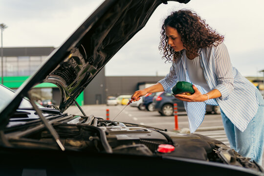 Young woman checking the oil level in her car engine using a dipstick, holding a bottle of motor oil, performing routine maintenance in a parking lot - Powered by Adobe