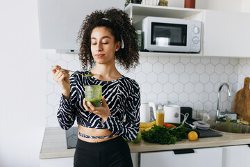 Athletic female with curly hair and dark skin enjoying smoothie, taking it with spoon from glass...