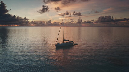 Fototapeta premium Sailboat resting on calm ocean waters of Bora Bora during a vibrant sunset, with colorful clouds reflecting on the water surface, creating a picturesque seascape. Exotic travel vacation getaway