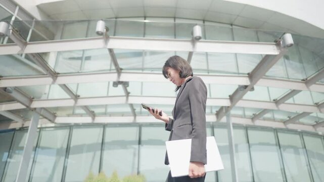 A woman is walking with a cell phone in her hand and a stack of papers. She is wearing a black suit and a black jacket