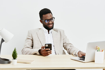 Businessman sitting at office desk using smartphone and working on laptop in modern workspace, professional male in glasses and beige blazer, workplace with plants and office supplies, concept of