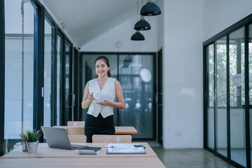 Asian businesswoman enjoying a coffee break in a modern office, standing beside a table filled with...