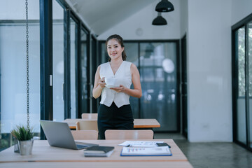 Young Asian businesswoman enjoying coffee during a break, standing next to a desk filled with a...