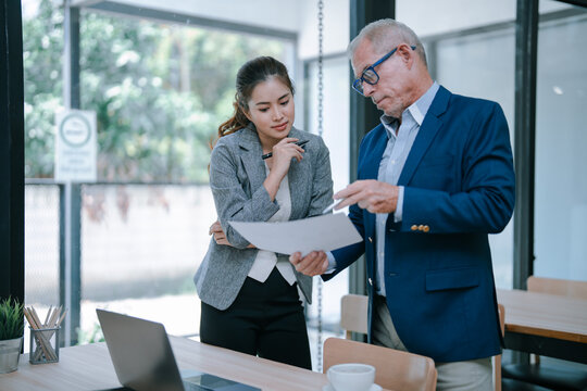 Senior manager pointing at a document and explaining contract details to a young businesswoman in a modern office, analyzing financial report, discussing project results or planning new strategy