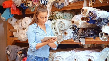 Young Woman With Digital Tablet Working In Fashion Design Studio Choosing Roll Of Fabric