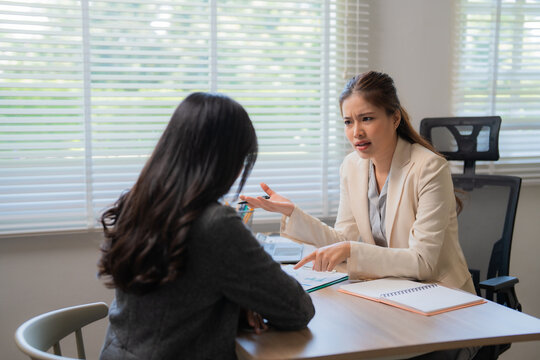 Two asian businesswomen are sitting at desk in the office and arguing about project, one of them is explaining something and pointing at documents while the other is listening