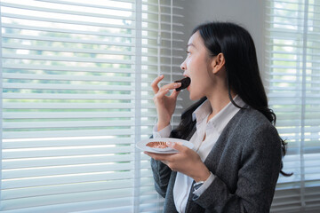 Obraz premium Asian businesswoman in smart casual outfit is eating a chocolate muffin during her office break, standing next to a window with venetian blinds, enjoying the view and the sweet treat