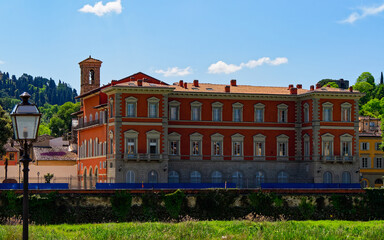 A red and white building with classical architecture,  arched windows and pilasters, situated alongside a grassy area. Its location is alongside the Lungarno, near the Arno River in Florence, Italy.