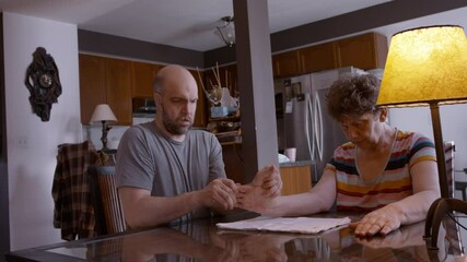 An adult son caregiver assists an elderly mother stroke survivor by gently stimulating and mobilizing her hemiplegic right thumb during a hand therapy session at the kitchen table, on cloth pad.