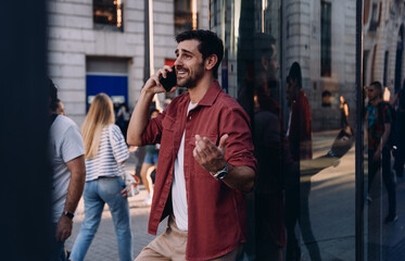 Obraz premium cheerful man in a red jacket leans against a glass wall, engaged in a phone call. Male reflection symbolizes digital communication, business networking, and modern technology in an urban setting.