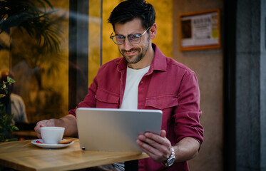 Male student studies online at a cozy café, deeply engaged with his tablet. Man balances education and lifestyle, using digital tools for learning while enjoying a warm coffee in an evening setting
