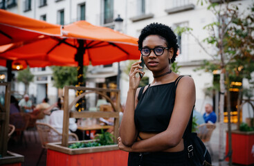 Trendy African American woman talking on smartphone outdoors near cozy street café, confidently looking at camera, wearing modern black outfit with hoop earrings and choker