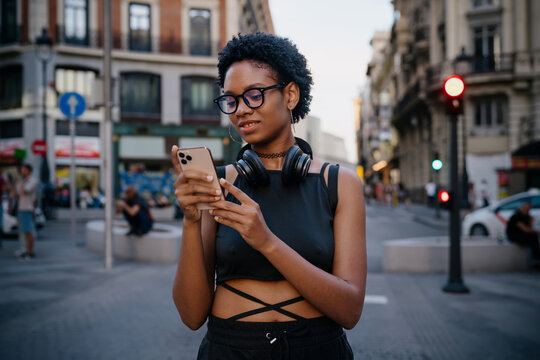 Young Black woman using smartphone on city street, wearing wireless headphones around her neck, checking messages or playlist with relaxed smile in urban daylight