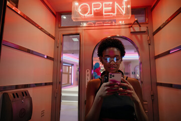 Young content creator standing beneath glowing “open” sign, focused on phone screen while...